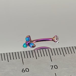 May include: A purple and blue titanium body jewelry barbell with a clear gem on one end and three blue opal gems on the other end. The barbell is measured against a ruler.