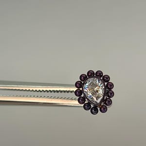 May include: A pear-shaped, clear gemstone surrounded by small, dark purple beads. The gemstone is held by a pair of silver tweezers, showcasing the intricate design and sparkle of the jewelry.