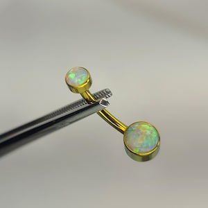 May include: A gold-colored belly button ring with two round opal stones. The opals have a mix of green, yellow, and white colors. The jewelry is held by a silver-colored tweezer.