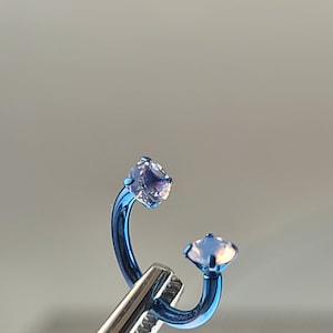 May include: A blue curved barbell body jewelry with two clear, iridescent gemstones. The jewelry is held by a silver tool against a neutral background. The gemstones have a square cut.