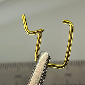 May include: A gold-colored metal wire bent into a U-shape with a smaller L-shaped wire attached to the top of the U-shape. The wire is held by a pair of pliers.