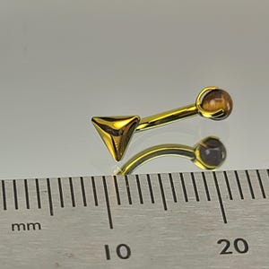 May include: A gold-colored eyebrow piercing with a triangular end and a rounded end with a brown stone. The curved barbell is approximately 0.75 inches long. The jewelry is displayed on a reflective surface.