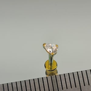 May include: A gold-colored labret stud with a clear, heart-shaped gemstone. The stud has a flat, circular base and a slender post. The gemstone is faceted, reflecting light. The background is a light gray, reflective surface.