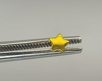 May include: A small, gold-colored star charm is held by a pair of silver tweezers. The star has a five-point design and a shiny, metallic finish. The tweezers are in sharp focus against a blurred background.