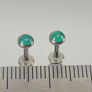 May include: Two body jewellery pieces with green gemstones, a silver post and a flat back. The jewellery is measured against a ruler.