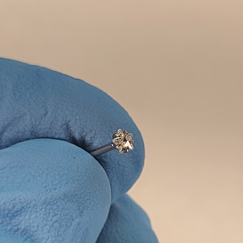 May include: A small, silver nose stud with a star-shaped, clear gemstone. The jewelry is held by a person wearing a blue glove. The stud's design features multiple facets, reflecting light.