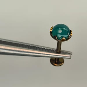 May include: A gold-toned labret stud featuring a smooth, round, teal-colored cabochon gemstone set in a scalloped prong setting. The jewelry piece has a flat circular back disc and is held by silver-colored metal tweezers for display.