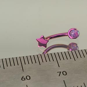 Peut inclure: Une barre incurvée en titane violette avec un charme en forme d'étoile et une pierre précieuse en opale. La barre est mesurée à l'aide d'une règle.