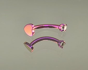 Può includere: Un paio di gioielli per il corpo in titanio viola con un'estremità a forma di cuore e un gioiello trasparente all'altra estremità.