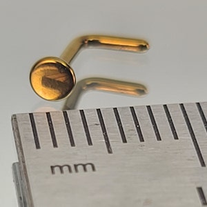 May include: A gold nose ring with a round bead at the end. The ring is measured against a ruler with millimeter markings.