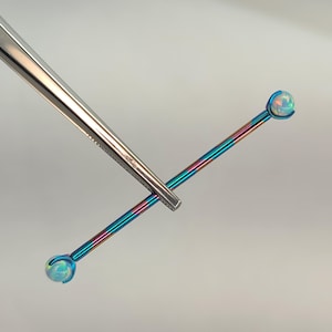 May include: A colorful industrial barbell piercing with opal-like ends. The barbell is held by silver tweezers. The barbell has a rainbow finish with blue, pink, and purple hues. The opal-like ends are round and have a pearlescent appearance.