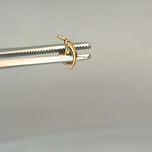 May include: A gold-colored, curved earring held by a silver surgical clamp. The earring has a smooth, polished finish and a crescent shape. The clamp's serrated jaws are visible, contrasting with the earring's shine. The background is a soft, neutral gray.