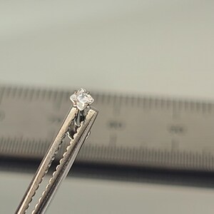May include: A small, square-cut, clear gemstone is held by a silver metal clip. The gemstone is centered in the frame, with a ruler in the background. The gemstone appears to be a diamond or cubic zirconia.