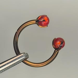 May include: A curved, bronze-colored body jewelry piece with a red, spherical gem at each end. The jewelry is held by a silver-colored tool. The gems have a translucent appearance, and the piece is set against a neutral background.