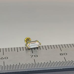 May include: A small, gold-colored stud earring with a rectangular, clear gemstone. The earring is positioned on a ruler, with the gemstone measuring approximately 0.3 inches in length. The earring has a screw-on backing.