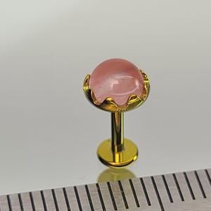 May include: A gold-tone labret stud featuring a smooth, round, pale pink cabochon stone set in a scalloped metal bezel. The jewelry is positioned next to a metal ruler, showing the stone has a diameter of approximately 0.25 inches.