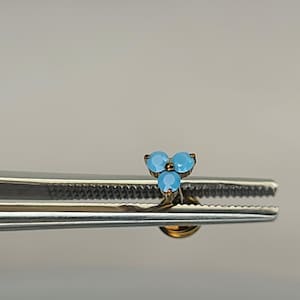 May include: A small, gold-toned earring featuring three light blue gemstones arranged in a triangular cluster. The earring is held by a silver-colored tool, showcasing its delicate design and color contrast against a neutral background.