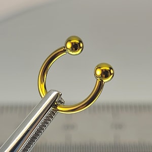 May include: A gold-colored horseshoe-shaped piercing with two small, round balls at each end. The piercing is held by a silver-colored tool, likely surgical forceps, against a light gray background.