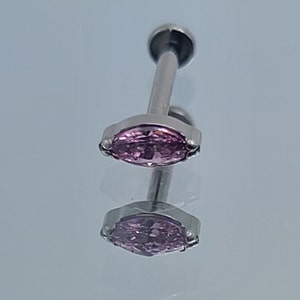 May include: A silver-colored, labret-style piercing stud with a marquise-shaped, light purple gemstone. The jewelry is displayed on a reflective surface, showcasing its design and color.