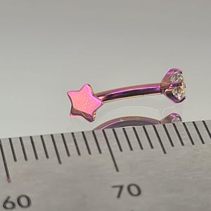 May include: Curved eyebrow ring with a star and a clear gem. The jewelry is a gradient of pink and purple. The star is on one end, and the gem is on the other. A ruler is in the foreground.