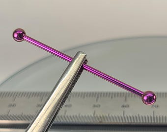 May include: A vibrant purple industrial barbell piercing. The barbell is held by silver tweezers, showcasing its straight bar design with a ball on each end. The piercing is set against a backdrop with measurement markings.