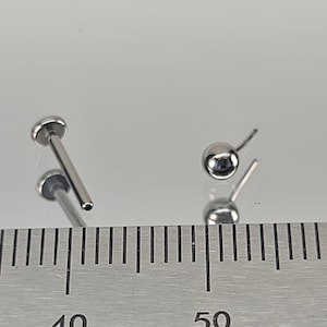 May include: Two silver body piercing studs, one with a ball end and one with a straight end, measured against a ruler.