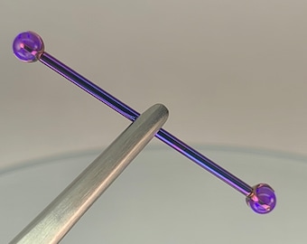 May include: A purple industrial barbell held by a silver tool. The barbell has a straight, slender bar with a spherical end on each side. The ends have a glossy, iridescent finish, reflecting shades of purple and blue.