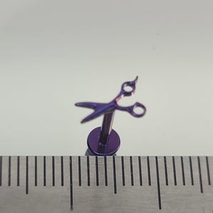 May include: A purple and blue scissor-shaped earring stud. The earring has a flat, circular base and a post. The scissors are detailed with open blades and finger holes. The earring is set against a white background.