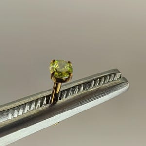 May include: A single, small, round, light green gemstone set in a gold-coloured metal setting. The gemstone is held by a pair of silver-coloured tweezers, with the gemstone and setting in sharp focus against a blurred background.