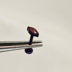 May include: A purple metal body jewelry stud featuring a marquise-cut dark red gemstone. The stud has a flat circular base and a thin post, held by metal tweezers to display the small, detailed construction of the piece.