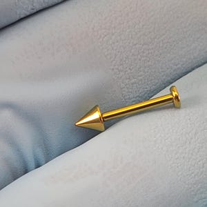 May include: Gold-colored body jewelry with a pointed tip. The jewelry is made of surgical steel and is suitable for lip, nose, or eyebrow piercings.