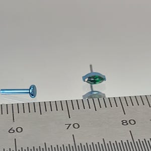 May include: Two small, light blue labret studs. One has a flat disc backing, the other a marquise-cut, emerald-green gemstone. The studs are shown next to a ruler for scale.