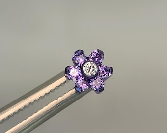 May include: A small, flower-shaped piece of jewelry with a central, clear stone surrounded by six purple stones. The jewelry is held by a silver tool against a gray background. The piece is likely an earring or body jewelry.