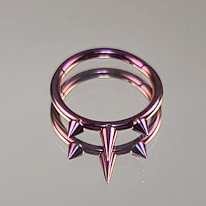 May include: A double-hoop septum ring with three pointed spikes. The ring is made of a rose gold colored metal.