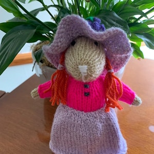May include: A knitted doll wearing a purple hat, pink shirt, and purple skirt. The doll has red yarn hair and a white face.