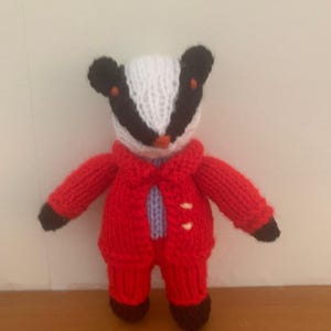 Puede incluir: Un muñeco de tejón tejido a mano con una chaqueta y pantalones rojos. El tejón tiene una cara blanca con marcas y orejas negras, y pies marrones. La chaqueta tiene dos botones blancos y una camisa azul claro debajo.