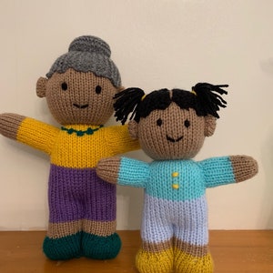 May include: Two knitted dolls, one with grey hair and a purple skirt, the other with black hair and a blue shirt, standing side by side on a wooden surface.