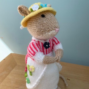 May include: A knitted mouse wearing a yellow hat with flowers, a pink and white striped sweater, and a white skirt with embroidered flowers.