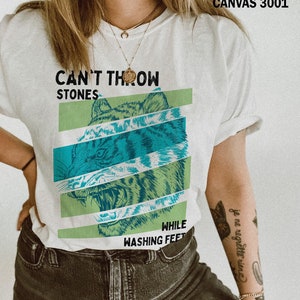 May include: White t-shirt with a green and blue tiger graphic and the text "Can't Throw Stones While Washing Feet".