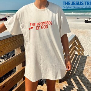 May include: A white t-shirt with the text "THE PROMISES OF GOD" in red lettering and two red hearts above the text. The t-shirt is being worn by a person standing on a wooden boardwalk.