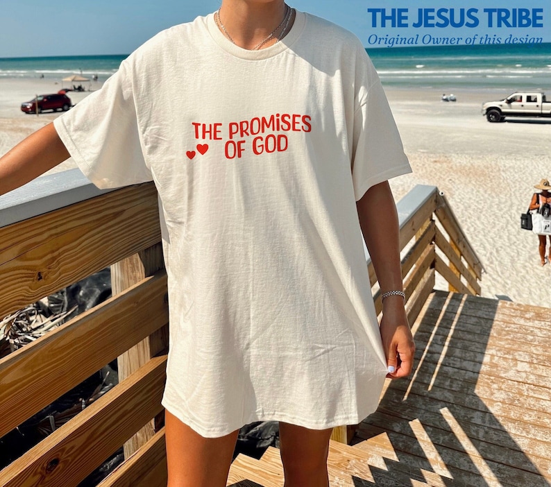 May include: A white t-shirt with the text "THE PROMISES OF GOD" in red lettering and two red hearts above the text. The t-shirt is being worn by a person standing on a wooden boardwalk.