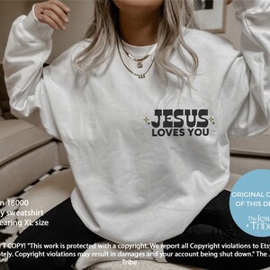 K&ouml;nnte beinhalten: Ein wei&szlig;es Gildan 18000 Sport Grey Sweatshirt mit dem Text "JESUS LOVES YOU" in Schwarz. Der Text wird von Stern-Grafiken begleitet. Das Sweatshirt ist ein l&auml;ssiges Kleidungsst&uuml;ck f&uuml;r den Alltag.