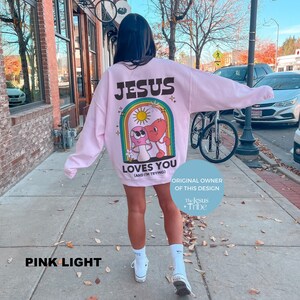K&ouml;nnte beinhalten: Hellrosa Sweatshirt mit dem Text "JESUS LOVES YOU (AND I'M TRYING)" und einer Grafik aus Pilz und Herz in einem Regenbogenbogen. Das Sweatshirt hat eine hellrosa Farbe.