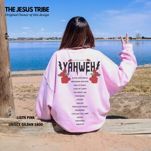 May include: A light pink sweatshirt with the text "YAHWEH" printed on the back. The text is surrounded by red roses and a lightning bolt. The text is also accompanied by a list of titles and descriptions, including "Alpha and Omega", "Beginning and End", "King of Kings", "Lord of Lords", "The Great I Am", "Firstborn", "Father", "Healer", "Prince of Peace", "Redeemer", "Lion and the Lamb", "Savior", "Shepherd", and "Creator".