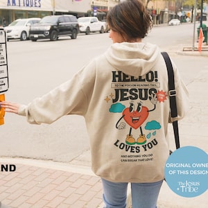 May include: A beige hoodie with a graphic print of a cartoon heart with eyes and legs. The heart is holding up two fingers in a peace sign. The text on the hoodie says "Hello! To the person reading this, Jesus loves you and nothing you do can break that love!" The text "The Jesus Tribe" is also visible on the image.