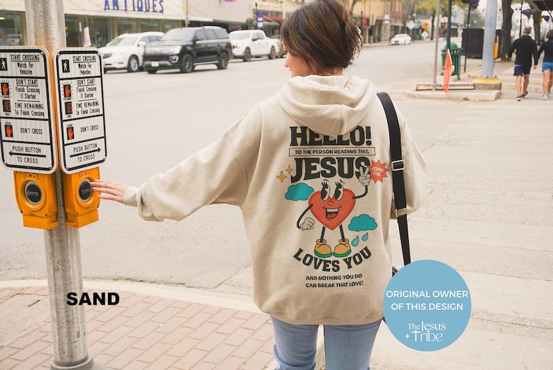May include: A beige hoodie with a graphic print of a cartoon heart with eyes and legs. The heart is holding up two fingers in a peace sign. The text on the hoodie says "Hello! To the person reading this, Jesus loves you and nothing you do can break that love!" The text "The Jesus Tribe" is also visible on the image.