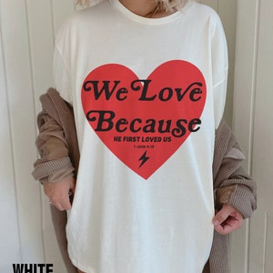 May include: White t-shirt with a large red heart graphic. The heart contains the words "We Love Because" and "He First Loved Us" with a lightning bolt. The word "WHITE" is at the bottom of the shirt.