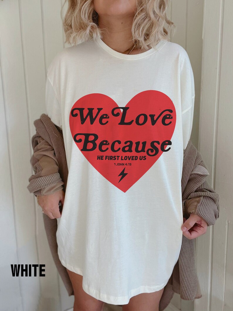 May include: White t-shirt with a large red heart graphic. The heart contains the words "We Love Because" and "He First Loved Us" with a lightning bolt. The word "WHITE" is at the bottom of the shirt.