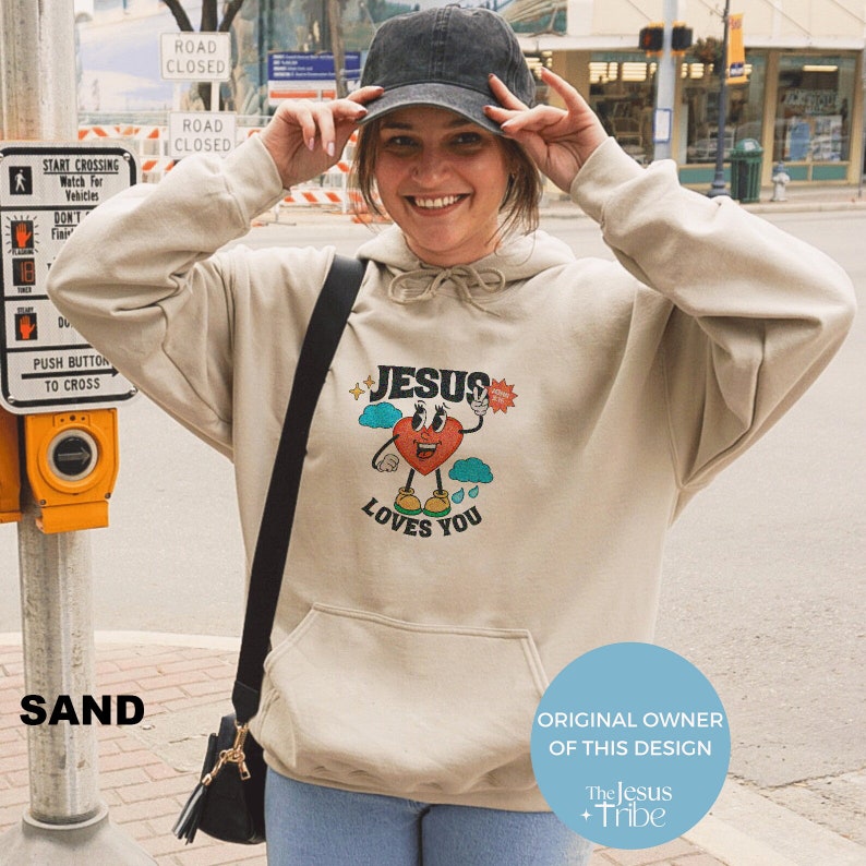 May include: A beige hoodie with a graphic print of a cartoon heart with a face and the words "Jesus Loves You". The hoodie has a hood and a kangaroo pocket. The graphic is in bright colors, including red, yellow, blue, and green.