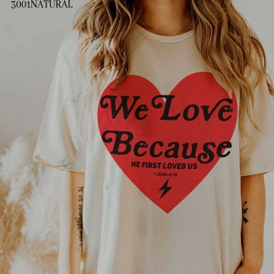 May include: Cream-colored t-shirt with a large red heart graphic. The heart contains the words "We Love Because" and "He First Loved Us 1 John 4:19" in black text, along with a lightning bolt. The shirt is made of Bella Canvas 3001 Natural material.
